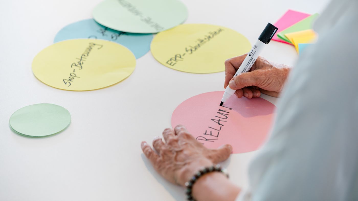 Eine Person schreibt mit einem Filzstift auf einen rosa Papierkreis. Weitere bunte Papierkreise liegen verstreut auf einem Tisch und deuten auf ein Brainstorming hin.